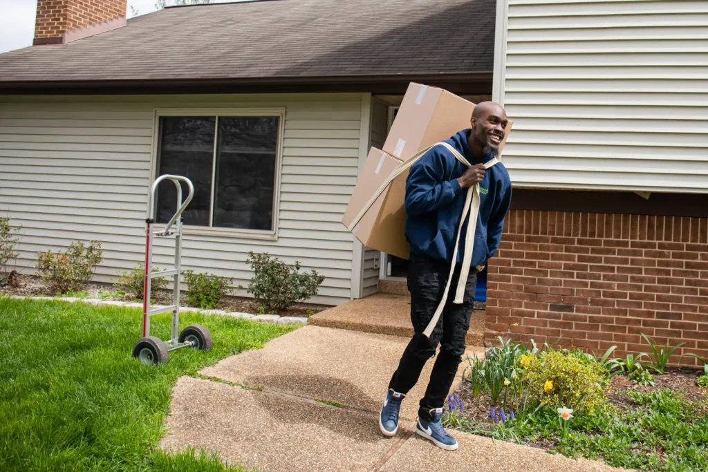 Smiling mover in a navy hoodie carrying stacked boxes on his back with a moving strap on a residential walkway.